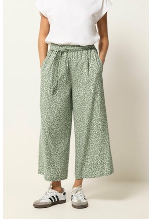 Osoba nosząca luźne, zielone spodnie z szerokimi nogawkami w kwiatowy wzór, związaną w talii, białą koszulkę i białe trampki z czarnymi paskami.