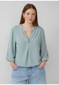 Hellgrüne Bluse mit langen Ärmeln, V-Ausschnitt, umgeschlagenen Bündchen und strukturierter Oberfläche, kombiniert mit hellblauen Jeans.