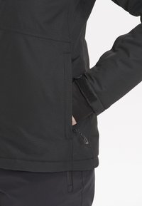 Schwarze wasserdichte Jacke mit strukturiertem Finish, ausgestattet mit einer seitlichen Reißverschlusstasche und verstellbaren Klettverschlüssen an den Bündchen. Schlichtes, funktionales Design.