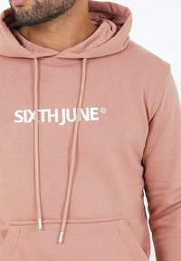 Uomo che indossa un hoodie rosa sporco con il testo bianco "SIXTH JUNE" e i lacci che pendono sul davanti.