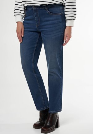 Vrouw die donkerblauwe rechte jeans draagt, zwarte enkellaarsjes met hak en een wit langärmelig shirt met zwarte horizontale strepen.