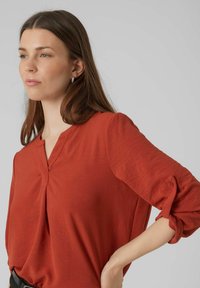 Blusa color ruggine con scollatura a V, maniche lunghe e polsini arrotolati. Tessuto morbido e testurizzato con una vestibilità rilassata. Design minimalista, senza motivi.