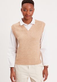 Chaleco de punto color marrón claro con escote en V y dobladillo acanalado, superpuesto a una camisa blanca con cuello, con textura suave y diseño ajustado.