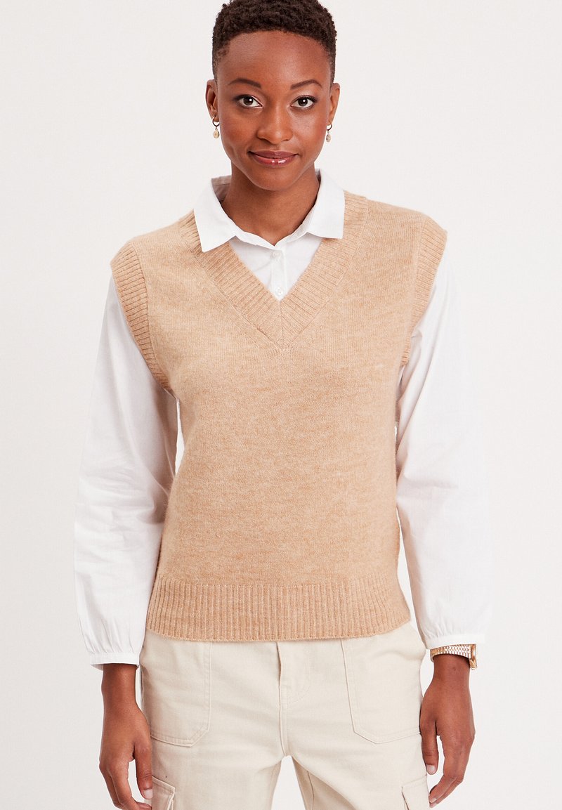Chaleco de punto color marrón claro con escote en V y dobladillo acanalado, superpuesto a una camisa blanca con cuello, con textura suave y diseño ajustado.