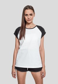 Camiseta blanca con mangas raglán negras. Material de algodón suave, corte holgado, cuello redondo y mangas cortas. Combinada con pantalones cortos negros desflecados.
