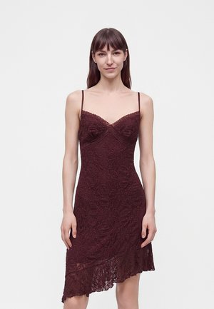 Jeune femme aux longs cheveux bruns porte une robe en dentelle rouge foncé sans manches avec de fines bretelles et un ourlet asymétrique sur un fond uni.