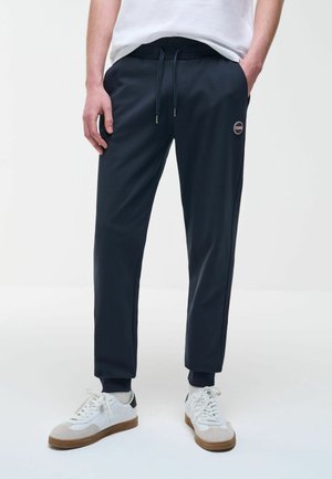 Persona che indossa pantaloni jogger neri con coulisse e sneakers bianche, con le mani nelle tasche, in piedi davanti a uno sfondo neutro.