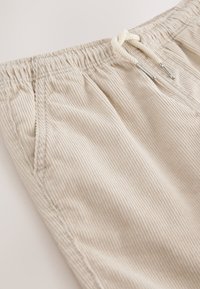Primer plano de pantalones de pana beige con cintura elástica, cordón con puntas metálicas y detalles de costura en los bolsillos laterales.