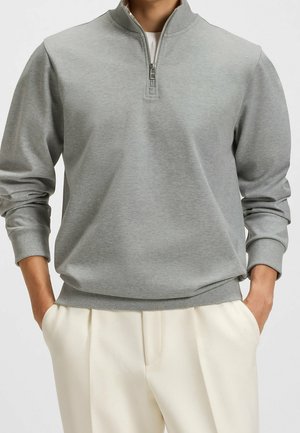 Man wearing a light grey quarter-zip pullover with long sleeves and cream-colored pants, hands in pockets, against a plain background.