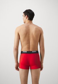 Boxer-culottes rouges en tissu lisse, avec une taille noire ornée du logo. Vue de l'arrière sur un fond uni.