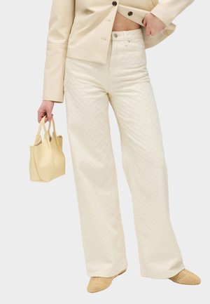 Kvinde iført cremefarvede bukser med vide ben, kort beige jakke, vævede beige sko, der holder en lille beige håndtaske mod en ensfarvet baggrund.