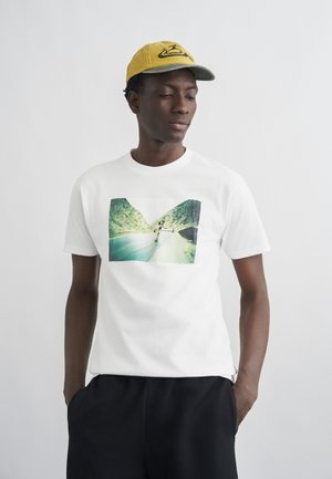 Άνδρας που φοράει κίτρινο καπέλο, λευκό μπλουζάκι με εκτύπωση φωτογραφίας σκέιτμπορντερ και μαύρο παντελόνι, στέκεται με τα χέρια στις τσέπες μπροστά σε λευκό φόντο.