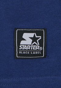 Kék anyag, amelyen egy fekete háttérrel rendelkező fehér "S" logóval és "STARTER BLACK LABEL" felirattal ellátott folt látható, fehér varrással keretezve.