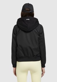 Czarna kurtka bomberka z kapturem, o gładkim wykończeniu, z elastycznymi mankietami i pasem, pokazana od tyłu. Połączona z jasnosłomkowymi legginsami.