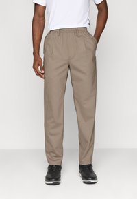 Pantaloni bej pentru bărbați, cu talie elastică, plisse în față și buzunare laterale, asortați cu pantofi negri cu șireturi.