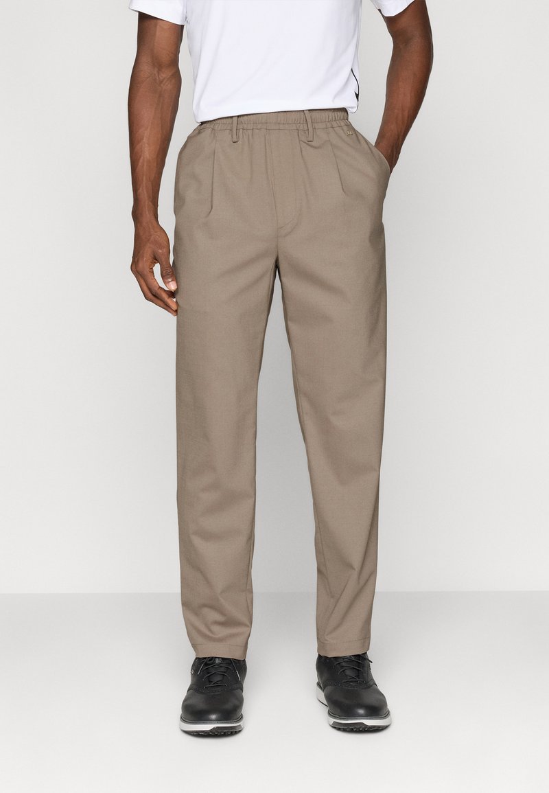 Pantaloni bej pentru bărbați, cu talie elastică, plisse în față și buzunare laterale, asortați cu pantofi negri cu șireturi.