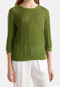 Grön stickad tröja med ribbat mönster, med trekvartsärmar, rund halsringning och en genomskinlig textur. Matchad med vita byxor.