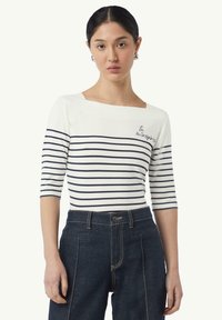 Gestreepte bateau-nekte top in wit en marineblauw, met 3/4 mouwen, met geborduurde tekst "Tu m’inspires," gecombineerd met donker denim hoge taillebroeken.