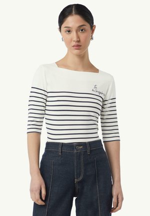 Gestreepte bateau-nekte top in wit en marineblauw, met 3/4 mouwen, met geborduurde tekst "Tu m’inspires," gecombineerd met donker denim hoge taillebroeken.