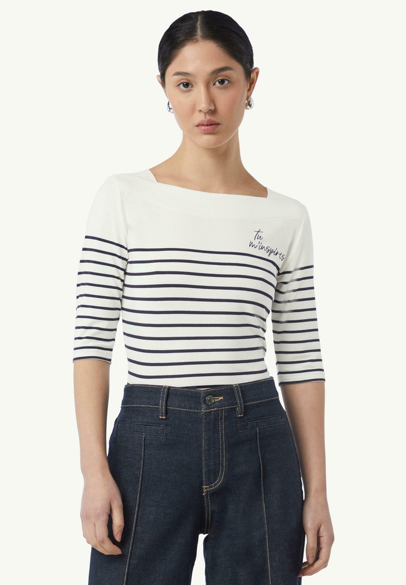 Gestreepte bateau-nekte top in wit en marineblauw, met 3/4 mouwen, met geborduurde tekst "Tu m’inspires," gecombineerd met donker denim hoge taillebroeken.