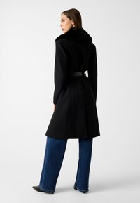Schwarzer Mantel mit einem Pelzkragen, tailliertem Bund und strukturiertem Design. Kombiniert mit weit geschnittenen blauen Jeans und spitzen schwarzen Schuhen.