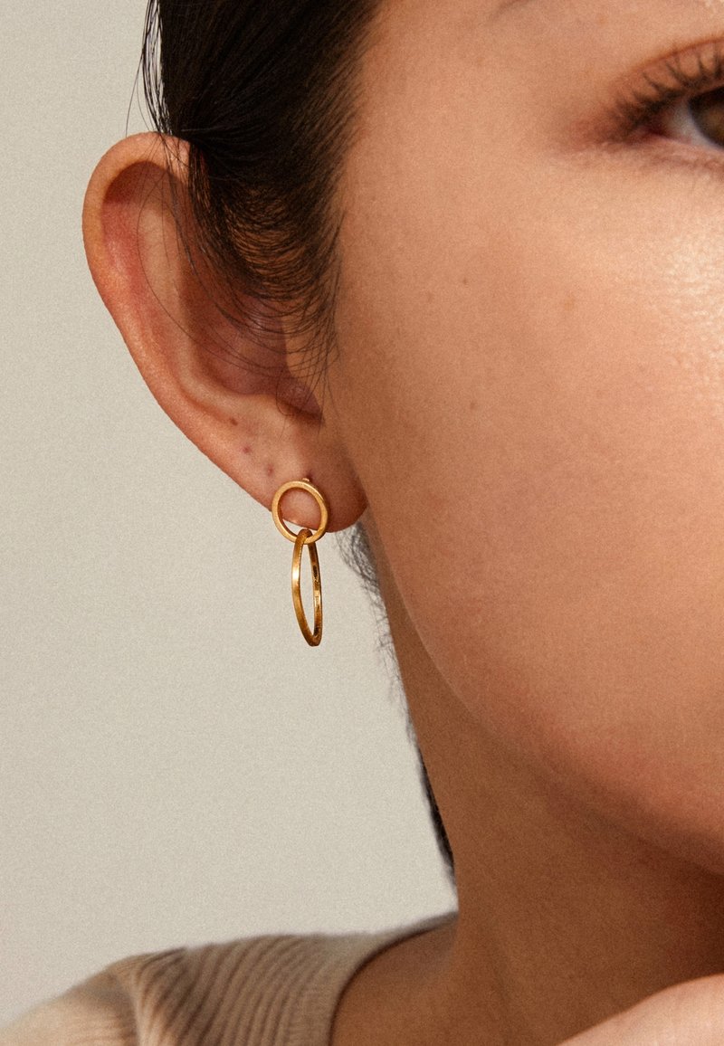 Gros plan sur l’oreille d’une femme portant une boucle d’oreille double en or sur un fond neutre.