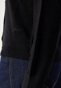 Pull noir en maille avec ourlet et poignets côtelés, présentant un logo de marque discrètement brodé près de la taille, porté avec un jean denim bleu foncé.