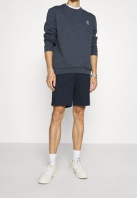 Persona che indossa un sudario a maniche lunghe grigio scuro, pantaloni corti blu navy, calze bianche, sneakers bianche e un orologio da polso argento su uno sfondo semplice.