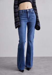 Siniset farkut, joissa on levenevät lahkeet ja korkea vyötärö. Perinteinen viisi-taskun muotoilu, sävyävää ompelua ja hieman joustavaa kangasta mukavuutta varten.