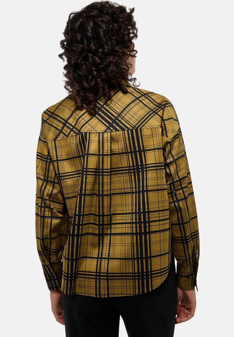 Chemise à manches longues en jaune moutarde avec un motif à carreaux noir, dotée d'un ourlet incurvé et d'un détail de dos avec empiècement. Texture de tissu lisse.