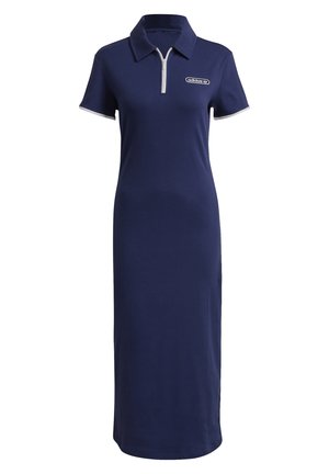 Femme portant une robe polo bleu marine à manches courtes avec fermeture éclair, bordée de blanc, et des baskets blanches, debout les mains relâchées le long du corps.