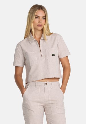 Kobieta o długich blond włosach, ubrana w beżowo-biały cienko w paski przycięty krótki rękaw i pasujące, wysokie spodnie z rękami w kieszeniach.