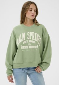 Jeune femme portant un sweatshirt vert clair "Palm Springs" et un jean bleu clair, se tenant devant un fond blanc uni.