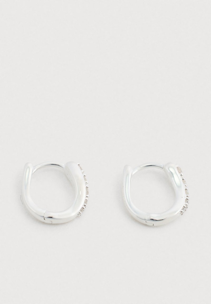 Cercei de tip cerc din argint cu finisaj lucios și formă curbată, sculpturală. O parte prezintă un rând de pietre mici integrate.