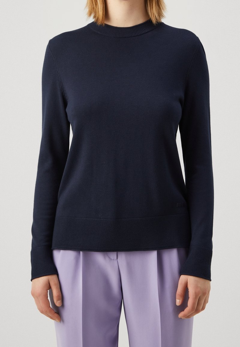 Femme portant un pull ajusté à manches longues bleu marine et un pantalon large lilas, debout devant un fond uni clair.