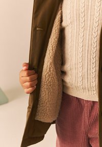 Khaki-jacka med en fluffig krämfärgad foder, kabelstickad krämfärgad tröja och rosa kordbyxor; hand som greppar jackans öppning.