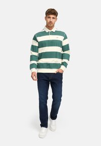 Camisa polo de manga larga con rayas horizontales verdes y blancas, cuello tipo polo y puños acanalados; combinada con jeans oscuros y zapatillas blancas.