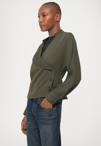 Top de color verde oliva, de manga larga, con un cuello doblado y detalles de botones en el dobladillo. Combinado con unos vaqueros azul oscuro, de corte clásico.