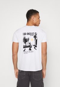 Biely bavlnený tričko s grafickým potlačou paliem, slnka a textom "LOS ANGELES KALIFORNIA" na chrbte. Krátke rukávy.