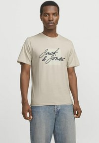 Beige katoenen t-shirt met zwarte cursieve tekst "Jack & Jones" en een verticale gestreepte achtergrond achter de tekst. Casual fit, ronde hals, korte mouwen.