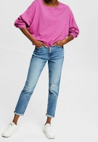 Różowy, oversize'owy sweter z podwiniętymi rękawami, w połączeniu z jasnoniebieskimi, postrzępionymi dżinsami i białymi sneakersami, prezentujący swobodny, luźny fason.