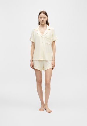 Jeune femme debout pieds nus portant une chemise crème à manches courtes boutonnée et un short assorti sur un fond blanc uni.