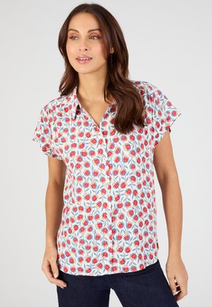 Donna che indossa una camicia bianca a maniche corte con bottoni e stampa floreale rossa, e jeans blu scuro, che guarda leggermente di lato.