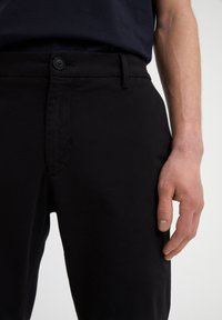 Man med svarta byxor och en marinblå skjorta, fokus på midjan, knappen, hällorna och den högra handen som vilar vid sidan.