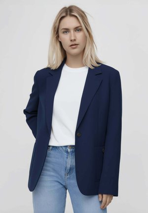 Giovane donna con capelli biondi indossa un blazer blu navy, una camicia bianca e jeans azzurri su uno sfondo chiaro e uniforme.