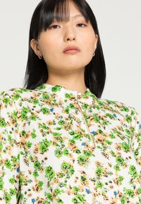 Blusa floral com base branca, apresentando padrões em verde, laranja e azul. Tem um decote alto e um detalhe de botão na frente.