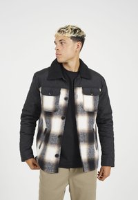 Kvadrovana črna jakna s karirastim vzorcem v beli, sivi in rjavi barvi. Ima ovratnik obložen s Sherpa materialom in dve žepe na prsih s gumbi.