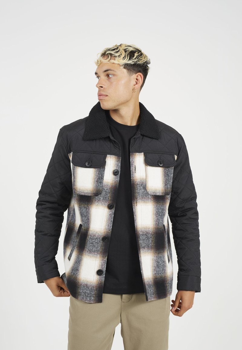 Kvadrovana črna jakna s karirastim vzorcem v beli, sivi in rjavi barvi. Ima ovratnik obložen s Sherpa materialom in dve žepe na prsih s gumbi.