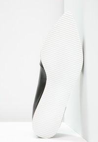 Czarne skórzane buty o gładkiej cholewce, spiczastym nosku i białej paskowanej gumowej podeszwie, ustawione pod kątem na jednolitym tle.