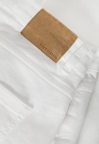 Pantalones de algodón blancos con una etiqueta de cuero que presenta el texto en relieve "TNTUUM". La costura visible y la textura suave realzan el diseño.
